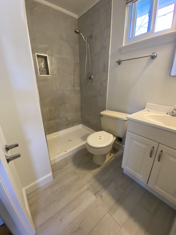 Finished bathroom with a grey stone-effect tiled shower, recessed niche, toilet and vanity under a window.