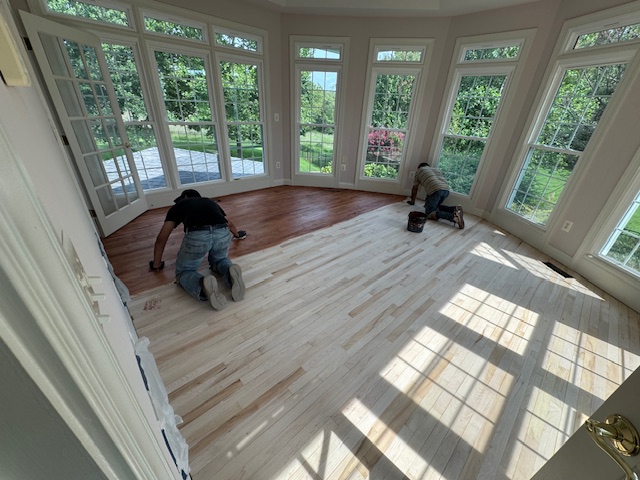 Worker sanding a hardwood floor in a bright sunroom lined with windows.