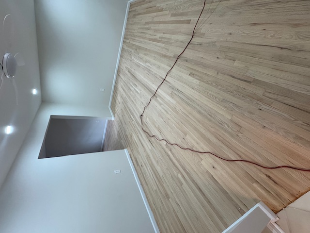 Long pale hardwood floor being sanded in a narrow hall with recessed lights.