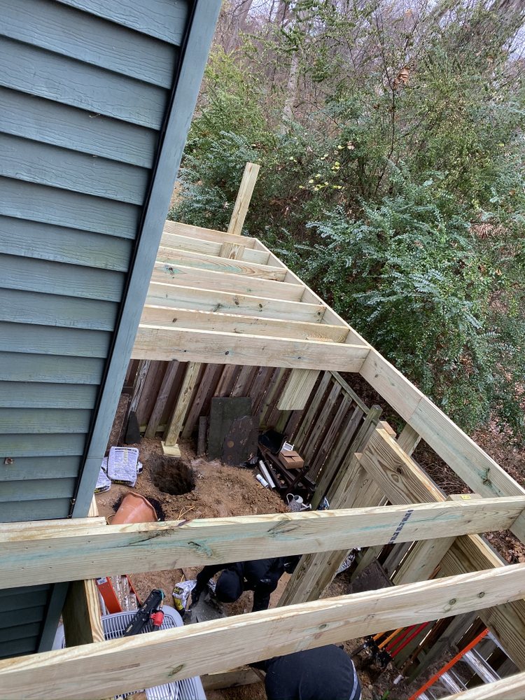 Crew working below new deck framing attached to blue lap siding on a hillside lot.